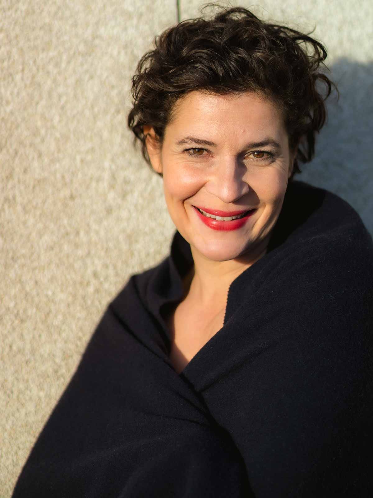 Cathérine Seifert lachend, in ein schwarzes Tuch gehüllt, an eine sonnenbeschienene Steinwand gelehnt.