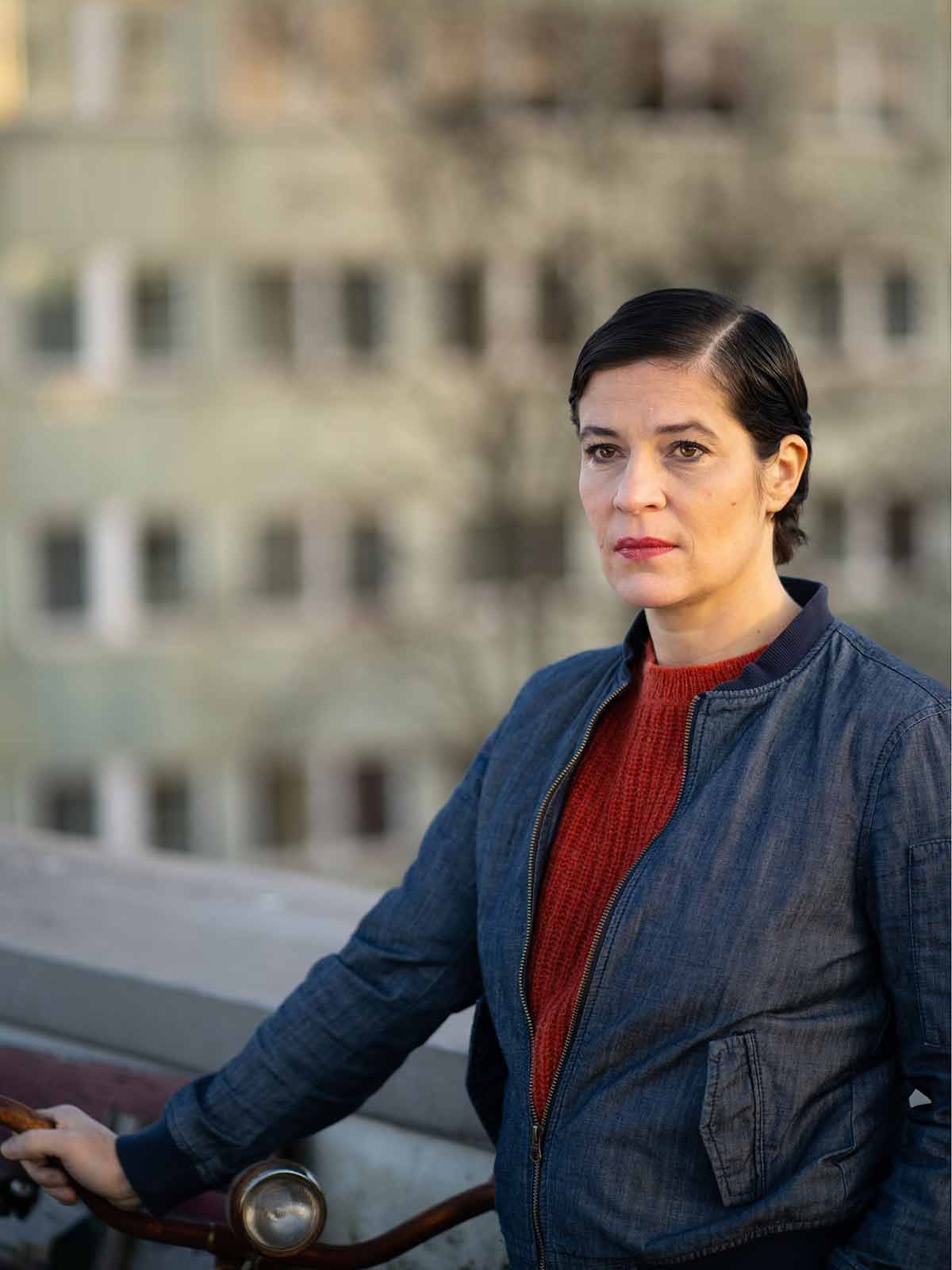 Cathérine Seifert in blauer Jacke über rotem Pullover, eine Hand am Lenker eines Fahrrads, im Hintergrund unscharf eine Hausfassade.